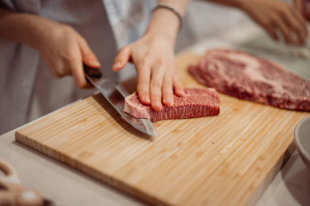 A pair of hands carefully slices marbled beef on a wooden cutting board, with another piece of raw meat in the background