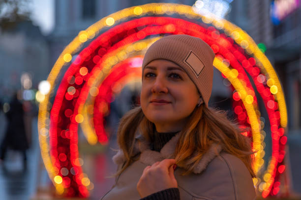 Portrait of serene adult caucasian woman in grey jacket and hat on evening winter city street. Blurred red and orange led illuminated Christmas bauble shaped decoration in the background. Soft focus.