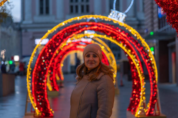 Portrait of serene adult caucasian woman in grey jacket and hat on evening winter city street. Blurred red and orange led illuminated Christmas bauble shaped decoration in the background. Soft focus.
