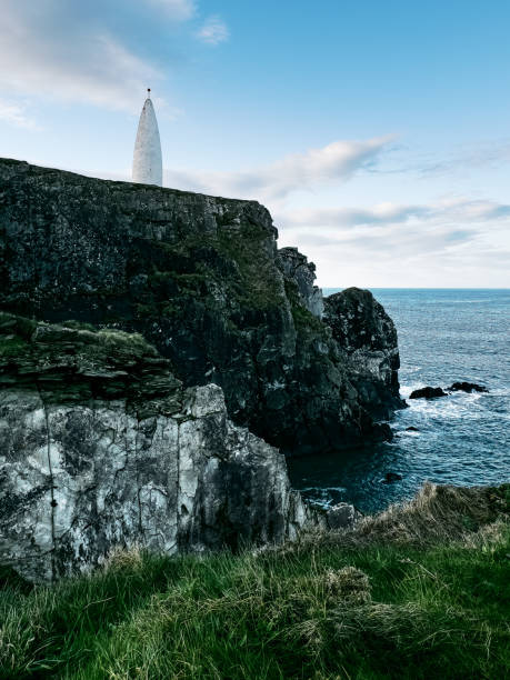 istock A lighthouse stands on a rocky cliff overlooking the sea, surrounded by grass. The sky is partly cloudy, adding a serene atmosphere to the coastal setting. 2186457618