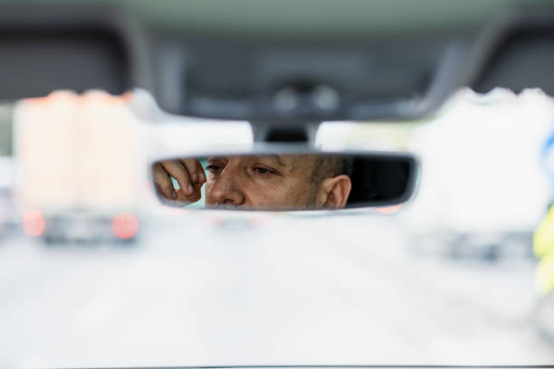 Mature man driving on a busy highway, adjusting his rearview mirror with a worried expression. Emotions of stress and frustration in daily commuting
