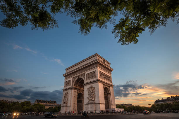 The majestic Arc de Triomphe in Paris during sunset, with a vibrant sky displaying hues of orange and blue. The monument is framed by leafy tree branches at the top. Cars and people are visible at the base of this famous landmark on the bustling Champs-Élysées.