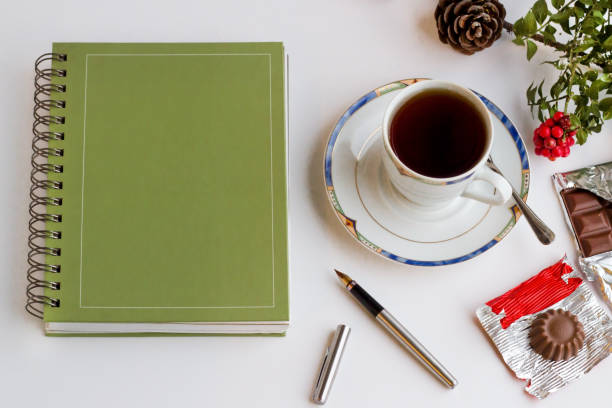 Above view of hard cover annual agenda on the white surface with stylo pen,a cup of tea and chocolate