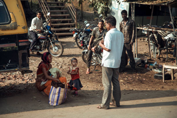 Paratwada Region, India - A young girl sits with a woman on the ground, while a man speaks to them in a bustling street filled with motorcycles and people during the day.
