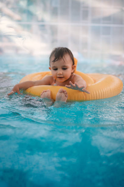 A happy toddler splashes in a pool while floating on a yellow inflatable ring and exploring his feet. The background features lush greenery, suggesting a scenic and relaxing outdoor environment. The child's expression captures pure joy and exploration.