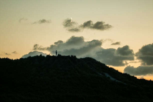On top of the dune looking over the sea during sunset on Terschelling. idyllic dune landscape on the Wadden island of Terschelling during sunset with the moon in the sky. In silhouette, a person stands on top of the dune looking over the sea with a crescent moon above it. Connection of man with nature and the insignificance of man. Philosophical image, time for resignation and reflection.