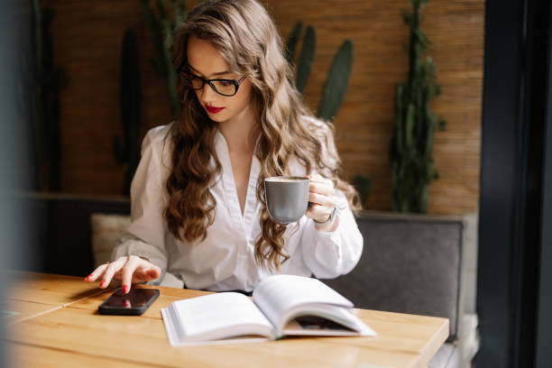Businesswoman drinking coffee and browsing smartphone while studying with open book at table in cafe