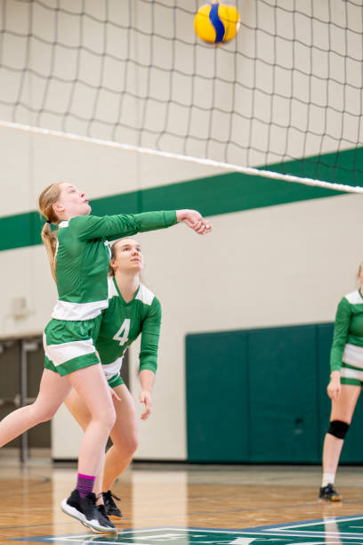 A group of women are seen playing a game of volleyball in a gym.  They are each wearing jerseys and are focused on setting up their plays as one player bumps the ball.