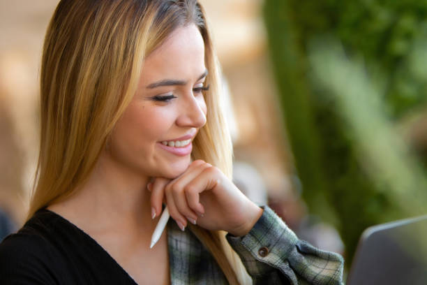 A beautiful young blonde woman, dressed in smart casual clothing, is working on a digital tablet with a digital pen in a modern coffee shop. Surrounded by lush green foliage, she is focused on reviewing financial data, embodying the entrepreneurial spirit of a small business owner. Her relaxed yet professional posture highlights the balance between leisure and productivity, as she seamlessly integrates work with her daily activities. This image is ideal for themes of entrepreneurship, small business management, and digital finance. Perfect for use in content related to business growth, online finance tools, or work-life balance in a modern, flexible workspace.