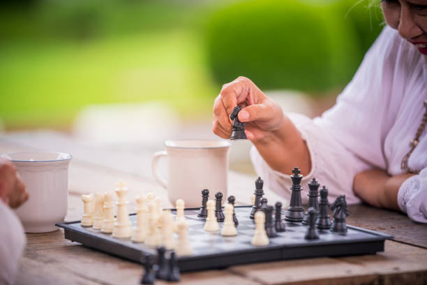 Two elderly women are sitting outdoors at a wooden table, playing chess holding a chess piece is time to relaxed of elderly relaxing on holiday lifestyle concept.