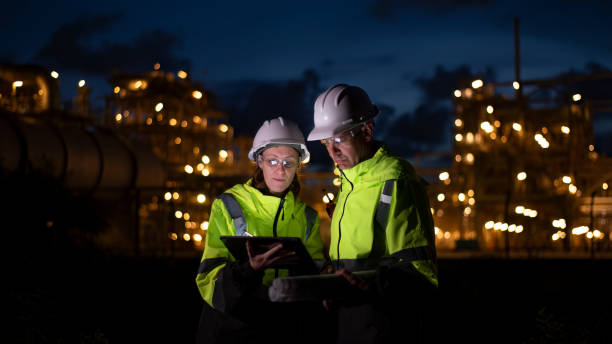 istock Engineers wearing safety gear, including hard hats examining survey are using tablet collaborating and discussing on screen work plant site use with night lights of oil refinery industry background 2186572483