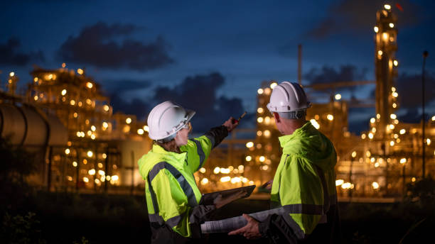 istock Engineers wearing safety gear, including hard hats examining survey are using tablet collaborating and discussing on screen work plant site use with night lights of oil refinery industry background 2186572484