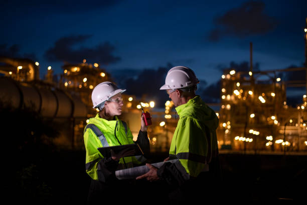 istock Engineers wearing safety gear, including hard hats examining survey are using tablet collaborating and discussing on screen work plant site use with night lights of oil refinery industry background 2186572485