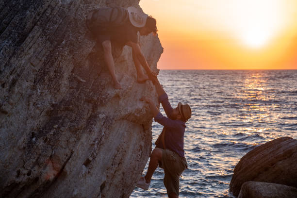 Two male hikers support each other climbing a rocky formation by the sea at sunset, showing teamwork, adventure, and friendship in nature