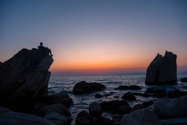 Silhouette of a young man reading a book while sitting on a rock overlooking a beautiful ocean sunset, creating a serene and inspiring scene