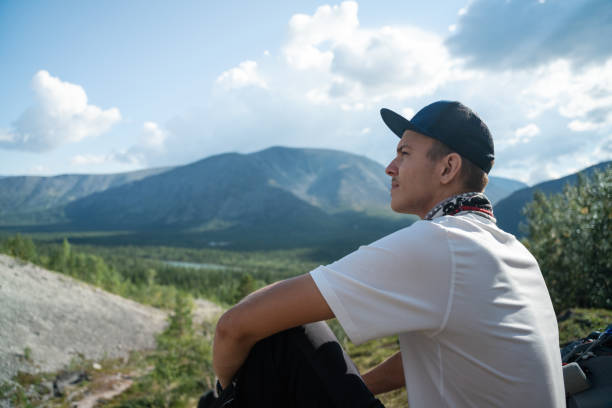 A young man in a cap and t-shirt sits peacefully, admiring a breathtaking mountain view under a clear sky. The serene outdoor scene embodies tranquility and the beauty of nature.