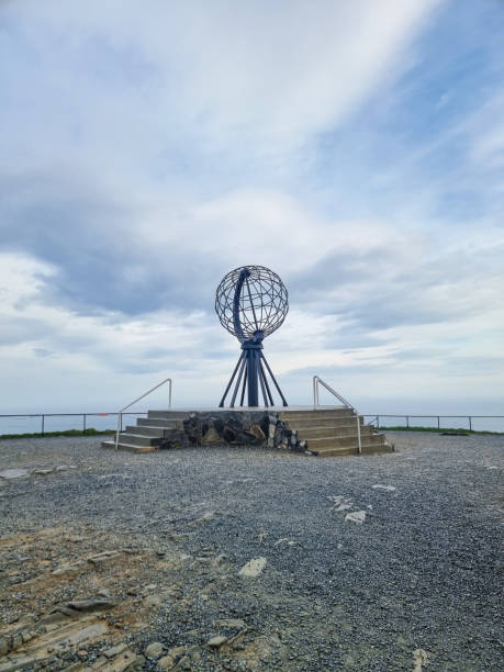 Visitors admire a striking globe monument at Nordkapp, standing proudly on rocky terrain, overlooking an expansive ocean. The sky is dotted with soft clouds, creating a tranquil atmosphere.
