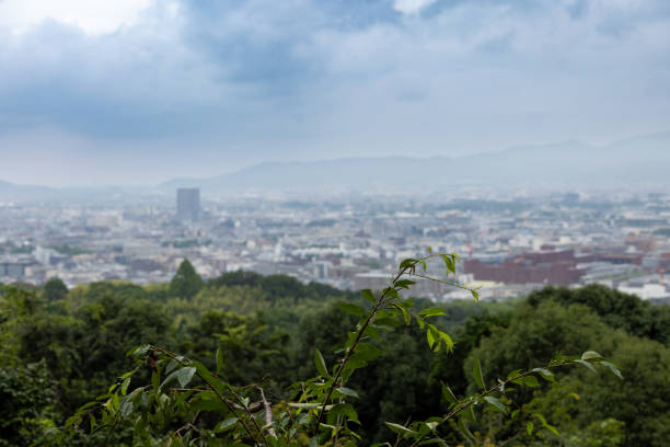 A captivating perspective of Kyoto, Japan, featuring the city’s expanse nestled among vibrant greenery in the foreground. The overcast sky creates a soft and tranquil atmosphere, highlighting the layers of modern architecture against the backdrop of distant misty hills. This shot beautifully encapsulates Kyoto’s harmony between its urban environment and lush natural surroundings.