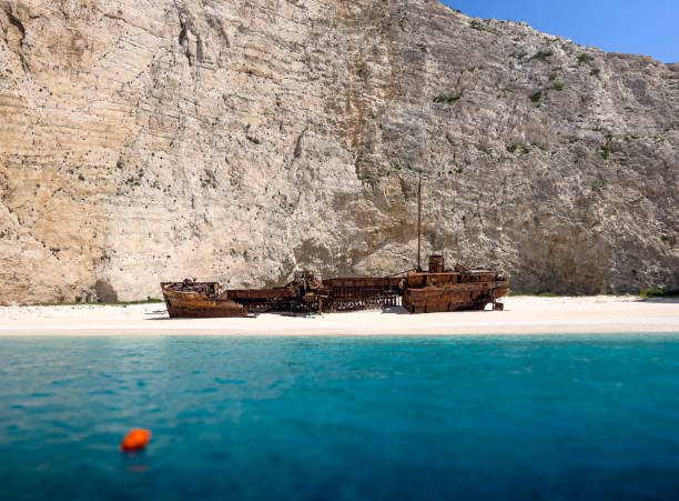 A rusty shipwreck rests on the white sand beach in Navagio Bay, Greece. The ship is partially submerged in the clear blue water, and the towering white cliffs of the bay rise in the background.