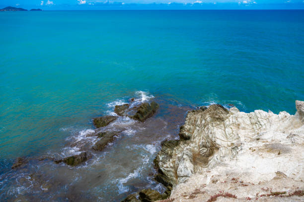 A rocky shoreline with a blue sky and ocean waves. The ocean is calm and the waves are small