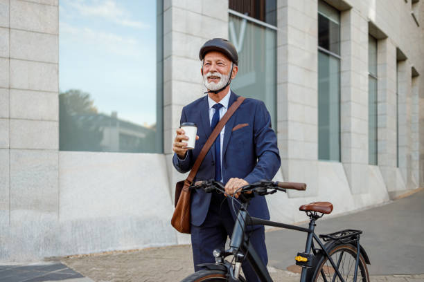 istock Confident elderly businessman with coffee cup and shoulder bag commuting with bicycle on city street 2186599846