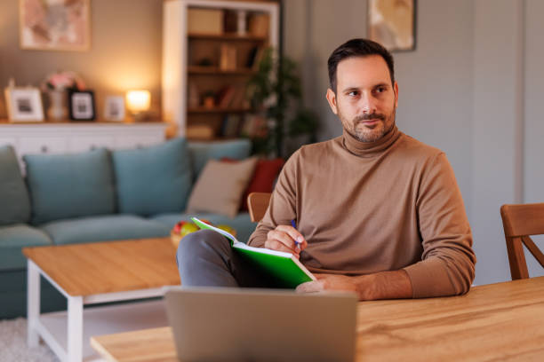 istock Thoughtful male freelancer looking away seriously while writing in note pad at desk in living room 2186599946