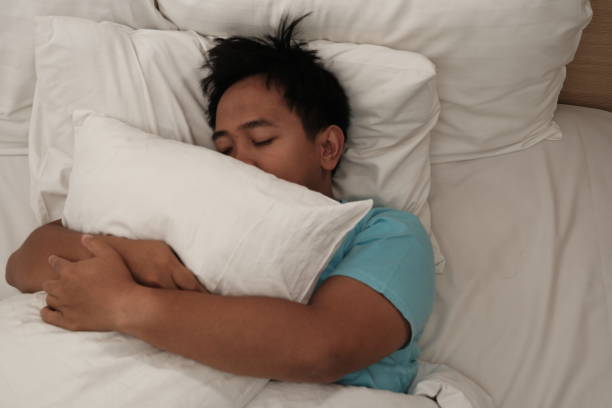 istock Asian man in a light blue shirt hugging a pillow while sleeping peacefully, surrounded by white bedding, creating a cozy and serene atmosphere. 2186609074