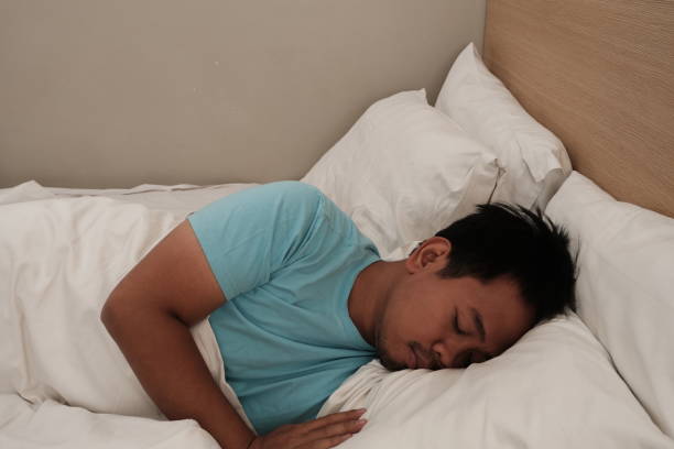 istock Asian man in a light blue shirt lying on their side, sleeping peacefully on a bed with white pillows and sheets, 2186609131