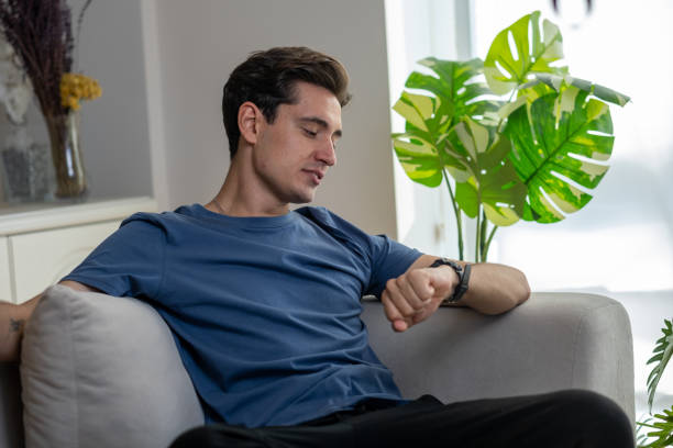 istock Young man sitting on sofa checking time on smartwatch 2186613391
