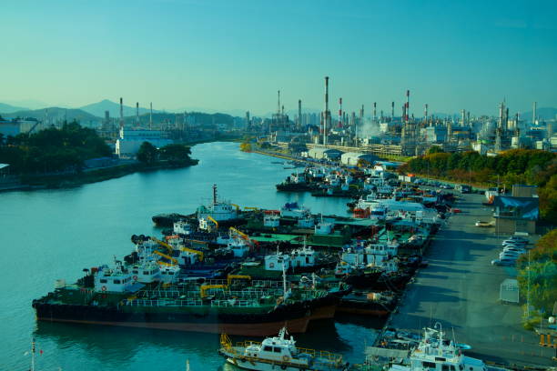 Ulsan, South Korea - November 14, 2024: A panoramic view of Jangsaengpo Port, showcasing docked vessels along the waterway with the towering chimneys of Yongyeon Industrial Complex in the background.
