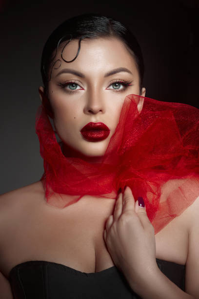 confident woman poses with striking makeup and bold red lips, dressed in a black outfit, while a flowing red scarf enhances her elegant appearance against a dark background.