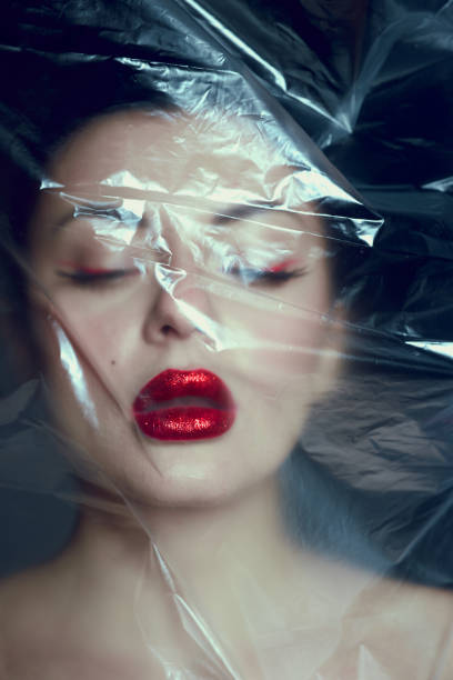 woman with bold red lips and artistic eye makeup poses with her face partially covered by translucent plastic wrap.
