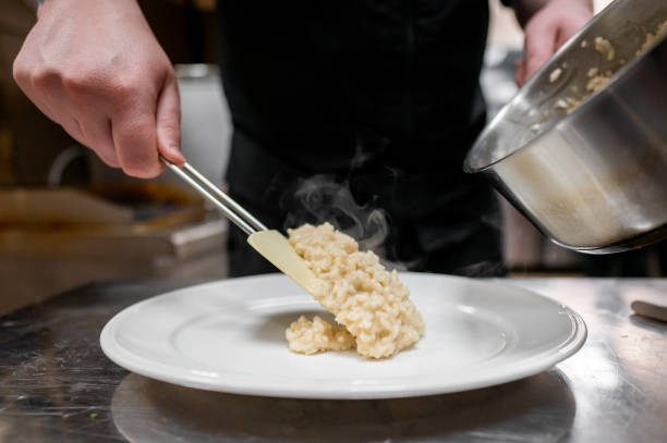 A chef gracefully plates creamy risotto from a pan onto a white dish, with steam rising, showcasing a refined culinary presentation in a professional kitchen setting.