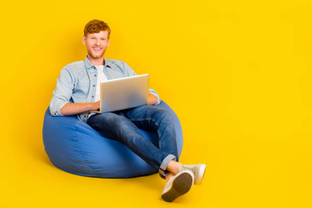 istock Full size body photo of young handsome chilling sitting pouf guy programmer using laptop distant work isolated on yellow color background 2186640852