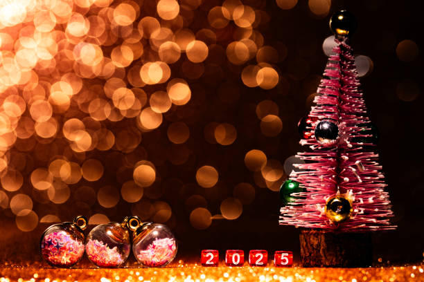 Charming New Year 2025 display featuring small red dice with white numbers forming 2025 set against a dark golden glitter background. On the left, four small star filled Christmas baubles add a festive touch, while a small pink Christmas tree stands to the right, bringing a playful and joyful element to the scene. Part of a series.