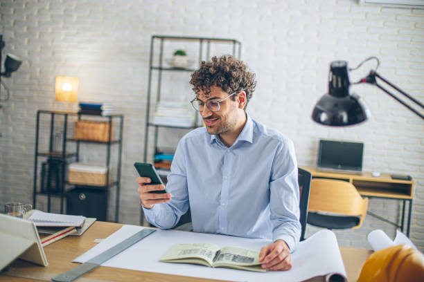 istock Focused architect using smartphone in bright modern office with blueprints on desk 2186666243