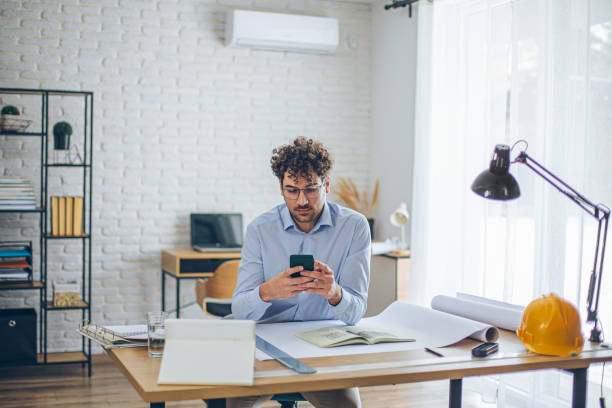 istock Focused architect using smartphone in bright modern office with blueprints on desk 2186666405