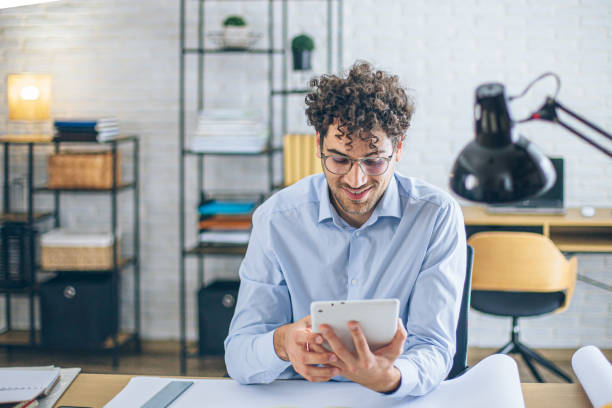 Focused architect using tablet in bright modern office with blueprints on desk