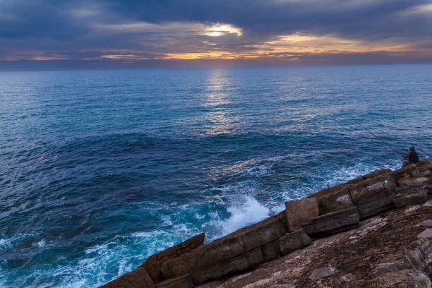 istock serene coastal scene during sunset. The ocean spans the majority of the frame, its surface glistening with reflections of the fading sunlight. Waves gently crash against rugged, textured rocks 2186674405