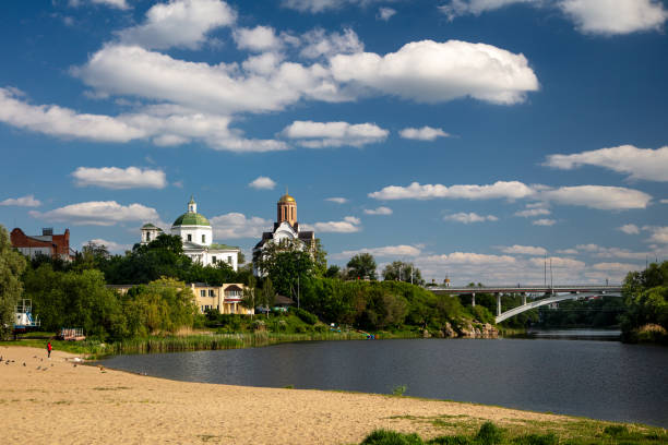 A quiet riverside location with a sandy beach, lush greenery and historic architecture with churches and cathedrals. The bridge spans the river under a bright blue sky with fluffy white clouds.
