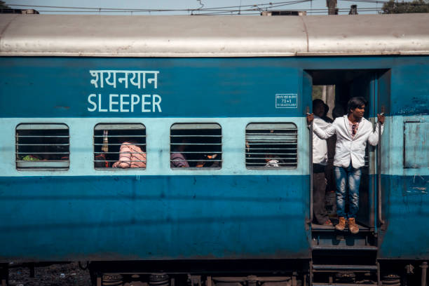 Maharashtra, India - A young man stands at the open door of a sleeper train, while others are seated inside. The vibrant, blue train shows signs of bustling activity typical of Indian rail journeys.