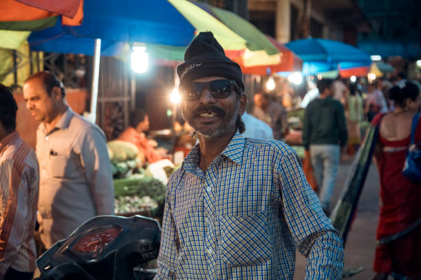 Pune Region, India - A man wearing a cap stands confidently in a vibrant market filled with colorful tents. Shoppers bustle around, enjoying the lively atmosphere as evening sets in.