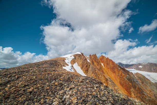 istock Snow-capped mountain ridge with cornice and red sharp rocks on top in sunlight. Snow cap and shiny pointy peak of gold color. Snow dome and colorful rocky peaked top. Sunny cloudy high mountains view. 2186697595