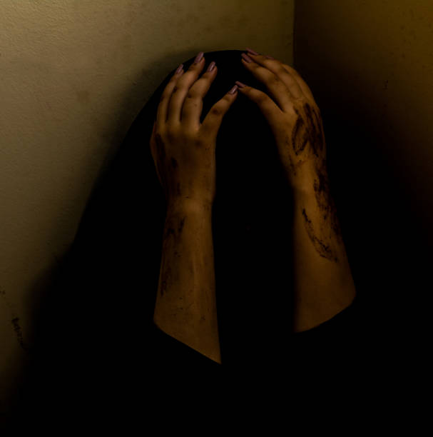 A dark and emotional scene of a woman sitting in a corner, covering her head with her hands, which are stained with smudges resembling dirt or paint. The dim lighting and shadows create a dramatic and moody atmosphere, evoking feelings of fear, distress, or solitude