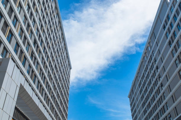 The image offers a perspective from the ground looking up towards the sky, framed by the towering facades of two modern high-rise buildings. The buildings, with their multitude of windows, stand tall against a backdrop of a clear blue sky speckled with a few wispy clouds. The architectural design is characterized by clean lines and a pattern of balconies that add a rhythmic visual element to the scene. The angle of the shot emphasizes the height and grandeur of the structures, while the sky provides a natural contrast to the man-made edifices. This urban landscape could be part of a bustling city's downtown core, where residential or commercial buildings reach towards the heavens, defining the city's skyline.