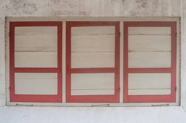 A vintage wooden window with three panels, featuring a red frame and beige shutters, set against a weathered wall. The rustic charm makes it ideal for architectural and historical themes