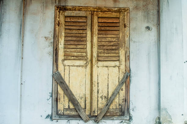 istock An old, weathered wooden window with shutters, secured by two diagonal planks, set against a worn, white wall. The rustic appearance suggests abandonment and history 2186736316