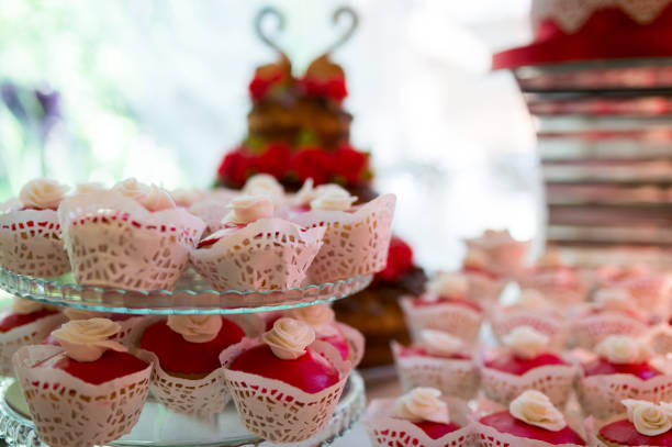 Close-up of a tiered glass stand with pink and white cupcakes topped with white frosting roses, arranged in a symmetrical pattern with more cupcakes in the background.