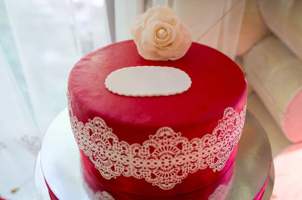 A birthday cake sits on a white plate with a lit candle on top. Red icing spells out 'Happy Birthday' surrounded by decorative icing and edible pearls, celebrating a special occasion.