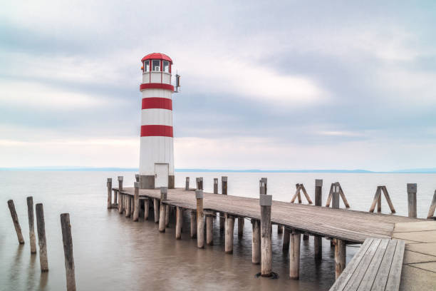 Red and white colored Lighthouse Podersdorf at the end of jetty going into the lake under the cloudy sky